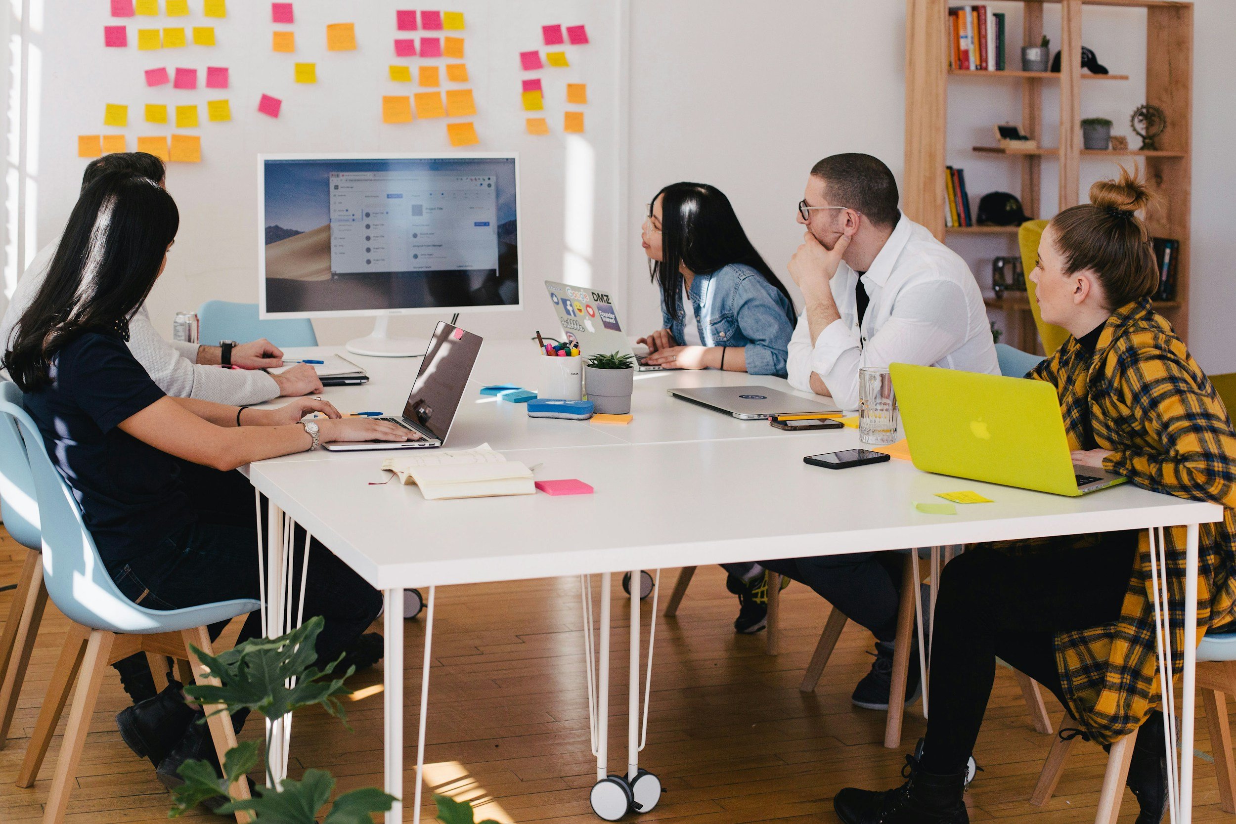 A group of five people are collaborating around a large white table in a modern office. Laptops and a desktop monitor are in use, and there are colorful sticky notes on the wall behind them.
