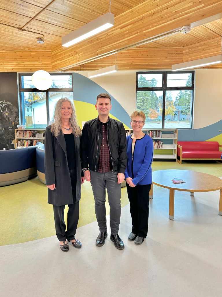 Robin, Graeme, and Sheila standing and smiling in a library with a wood ceiling and seating and shelving in the background