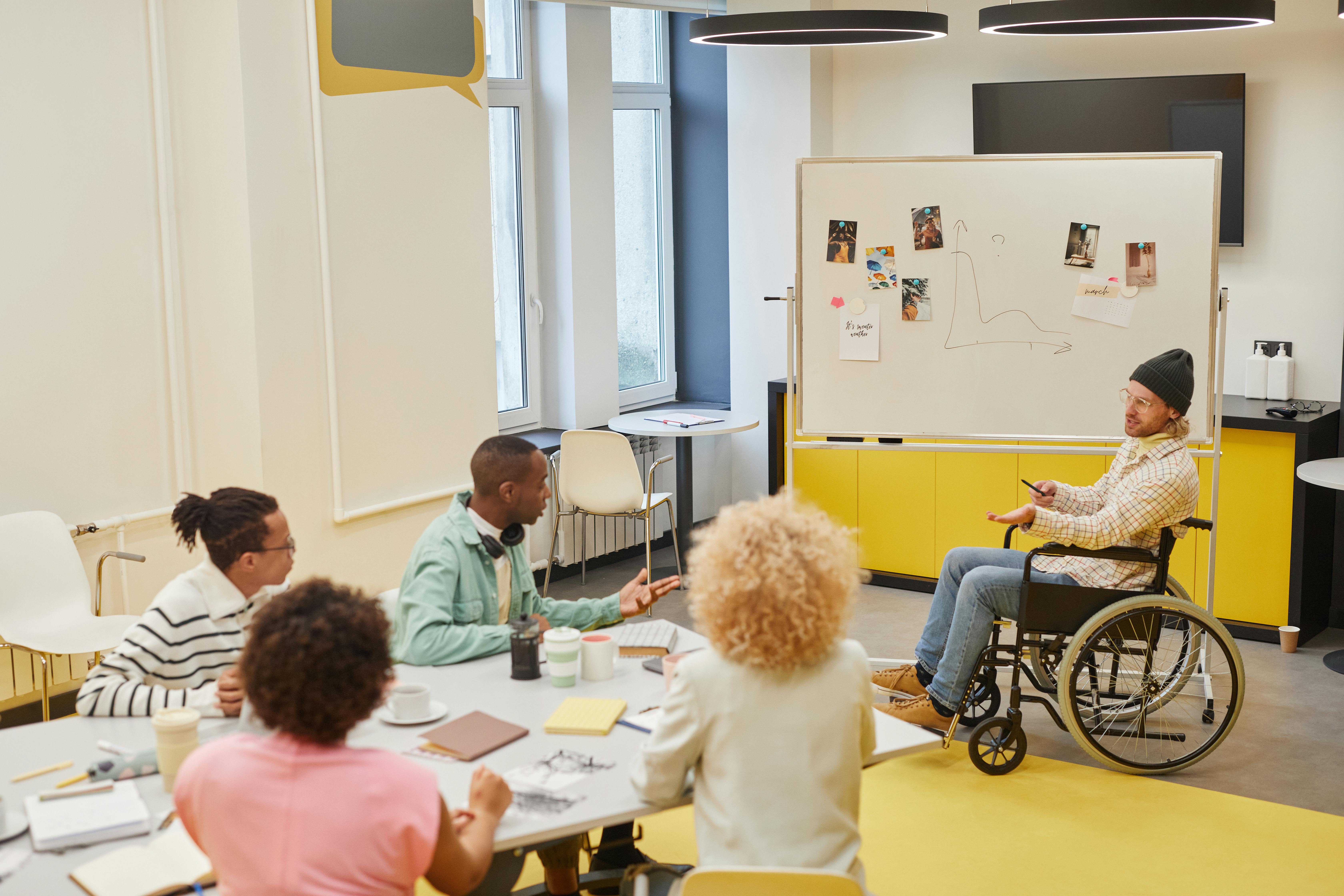 Four people, one of whom is in a wheelchair, are seated around a light-colored table in a spacious, modern meeting room. They appear to be engaged in a discussion or presentation, with a whiteboard visible in the background.
