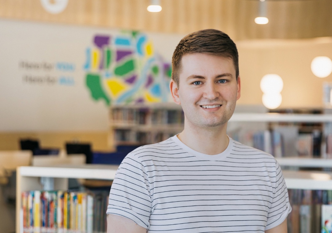 Headshot of Graeme standing in a brightly-lit library with bookshelves behind him